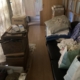 長年使用されていなかった木造平屋の廊下に、布団や段ボール、紙袋、雑貨類が無造作に積み上げられ、通路が塞がれている状態のゴミ屋敷。自然光が遮られ、生活動線が確保できないほど物が散乱している様子。