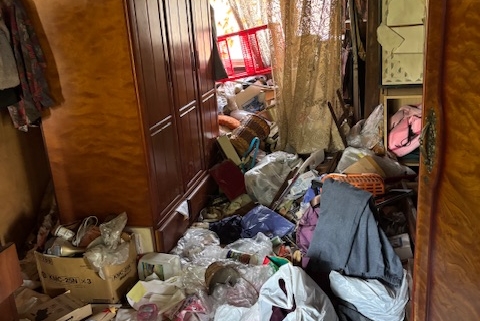 栃木県宇都宮市の6DK戸建て住宅の室内。衣類や生活用品、袋類が床一面から膝の高さ付近まで堆積し、通行が困難なゴミ屋敷状態。