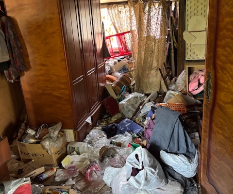 栃木県宇都宮市の6DK戸建て住宅の室内。衣類や生活用品、袋類が床一面から膝の高さ付近まで堆積し、通行が困難なゴミ屋敷状態。